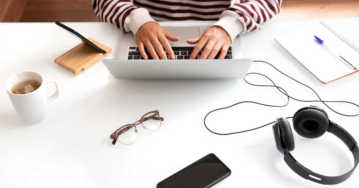 una persona lavora alla scrivania con il suo computer portatile, sul tavolo un cellulare, delle cuffie, un paio di occhiali